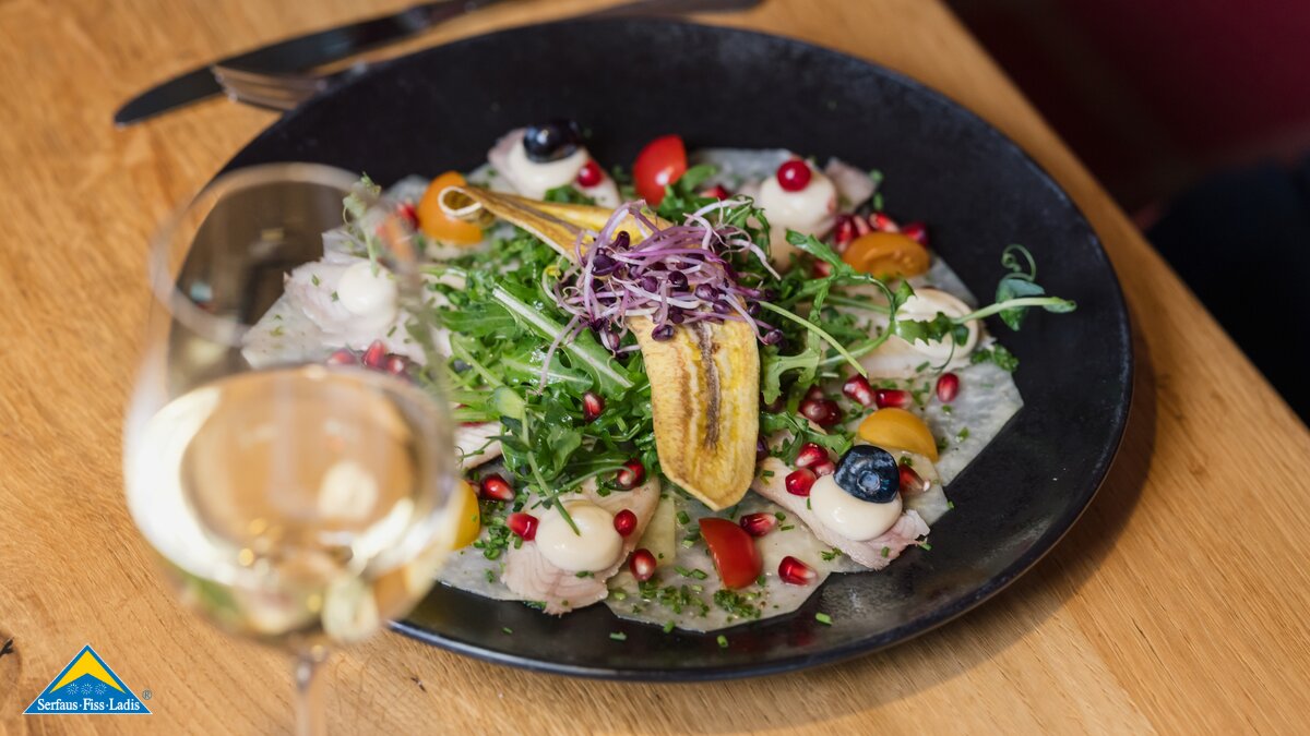 Ein eleganter Teller mit kreativ angerichtetem Salat, Granatapfelkernen, Früchten und einer knusprigen Bananenscheibe, daneben ein Glas Weißwein | © Serfaus-Fiss-Ladis Marketing GmbH | Fabian Schirgi