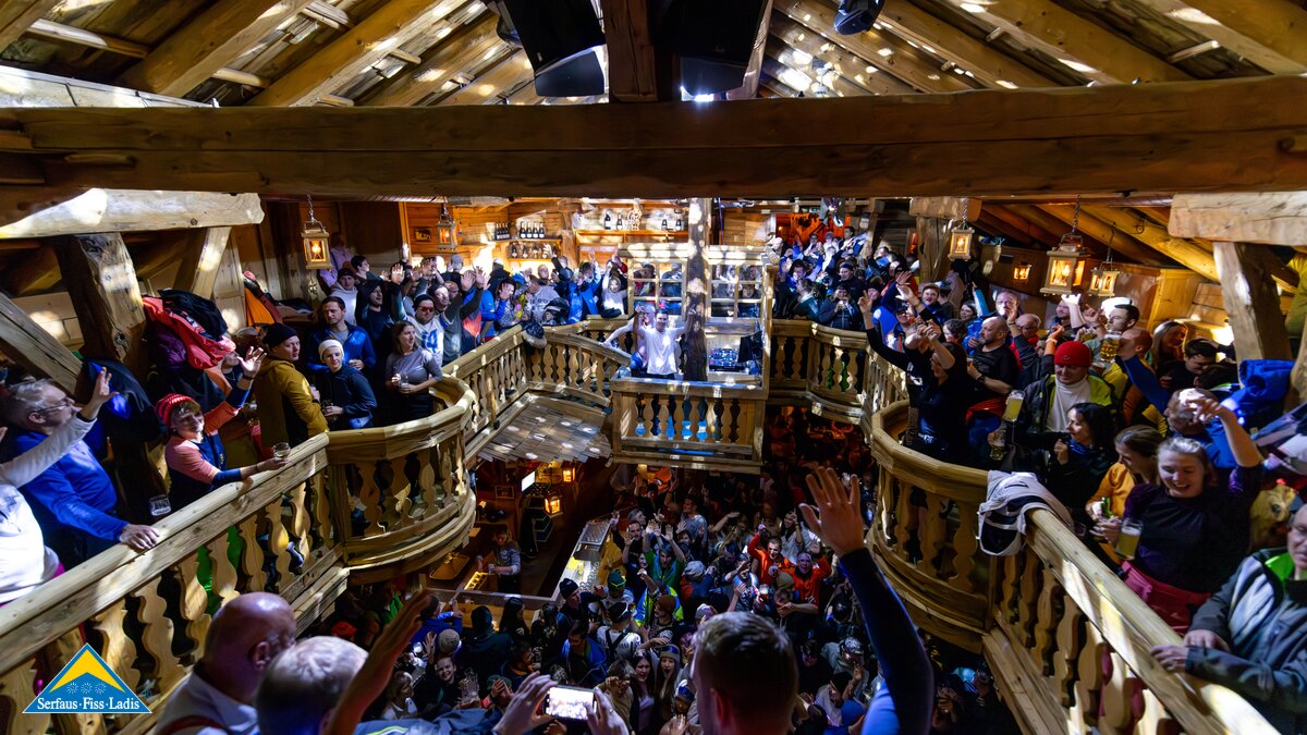 Menschenmenge feiert ausgelassen in einer gemütlichen, zweistöckigen Holzhütte mit Musik und Getränken | © Serfaus-Fiss-Ladis Marketing GmbH | Fabian Schirgi
