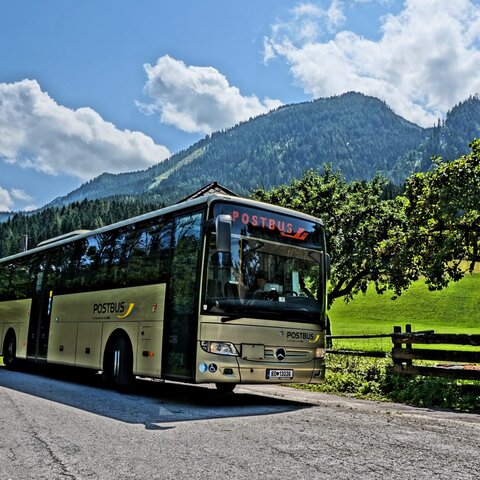 Ein gelber Postbus steht auf einer Landstraße, umgeben von Wiesen, Bäumen und Bergen unter blauem Himmel | © Serfaus-Fiss-Ladis Marketing GmbH