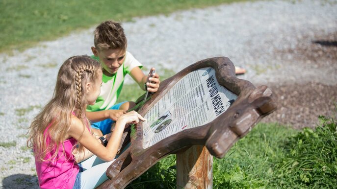 Zwei Kinder lesen gespannt eine kreativ gestaltete Informationstafel aus Holz für das Wo-Bä-Lu Geocaching in Serfaus-Fiss-Ladis | © Serfaus-Fiss-Ladis Marketing GmbH | Andreas Kirschner