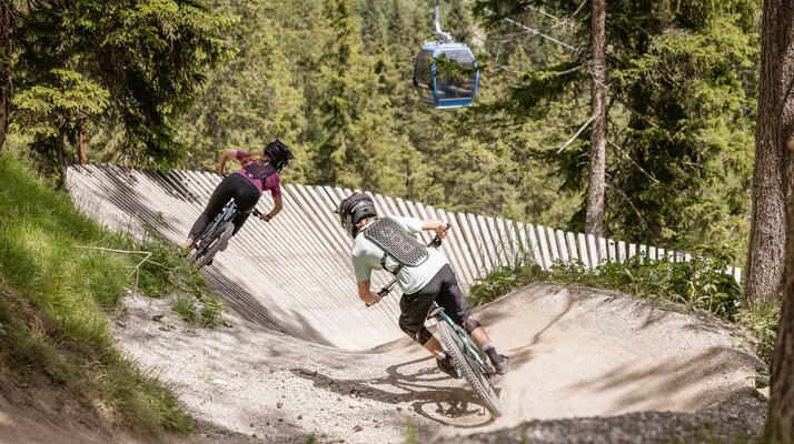 Zwei Mountainbiker fahren auf einem kurvigen Trail durch den Wald, eine Gondel schwebt darüber | © Serfaus-Fiss-Ladis Marketing GmbH