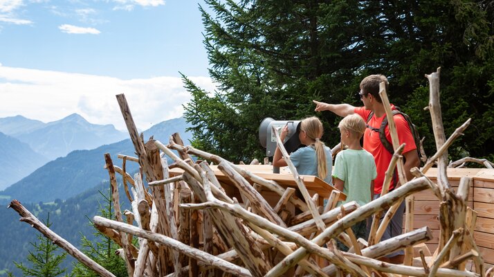 Familie beobachtet gemeinsam durch ein Fernrohr die alpine Landschaft auf dem Wildtierweg in Serfaus-Fiss-Ladis, umgeben von Holzskulpturen und Bäumen | © Serfaus-Fiss-Ladis Marketing GmbH | Andreas Kirschner