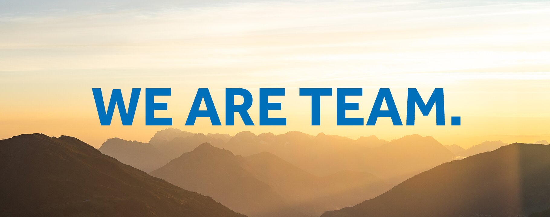 Sonnenaufgang über einer Berglandschaft mit dem zentralen Schriftzug „WE ARE TEAM.“ in blauer Schrift