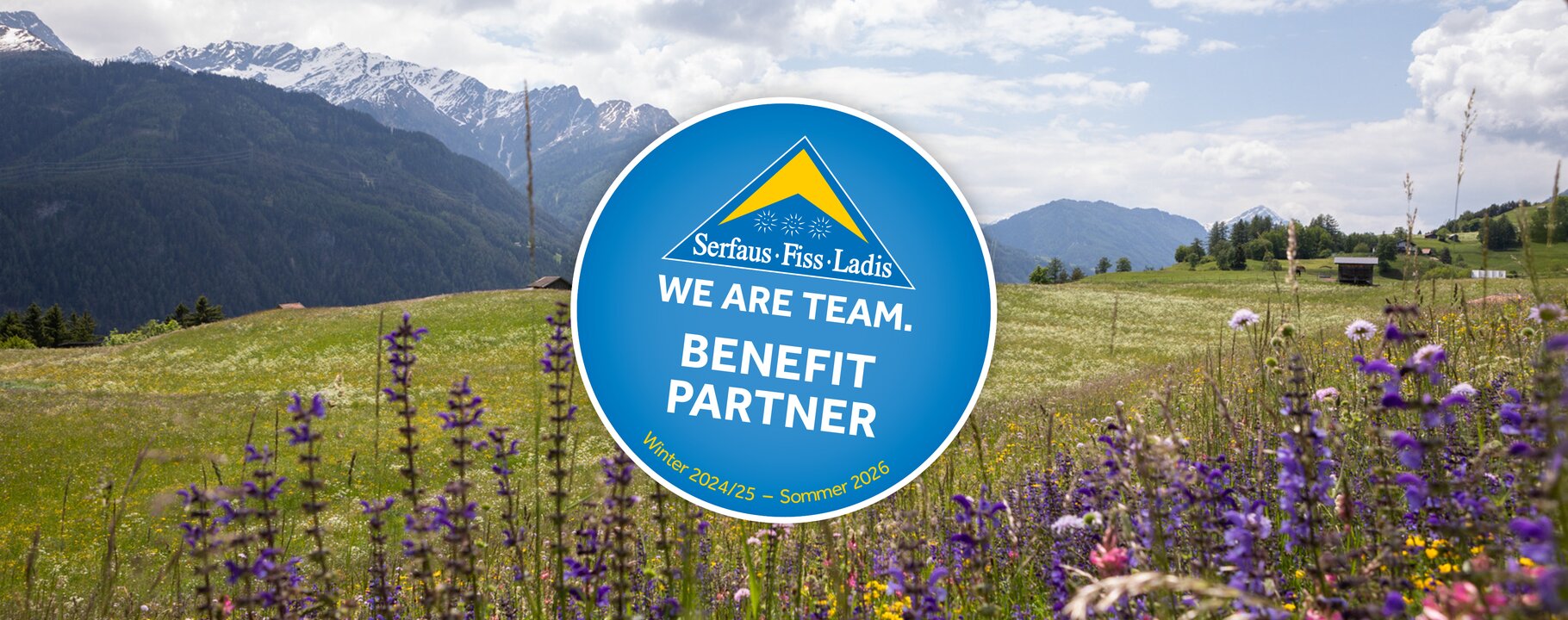 Rundes „WE ARE TEAM. BENEFIT PARTNER“-Logo in einer bunten Bergwiese mit Blick auf die Tiroler Alpen und Serfaus-Fiss-Ladis | © Serfaus-Fiss-Ladis Marketing GmbH