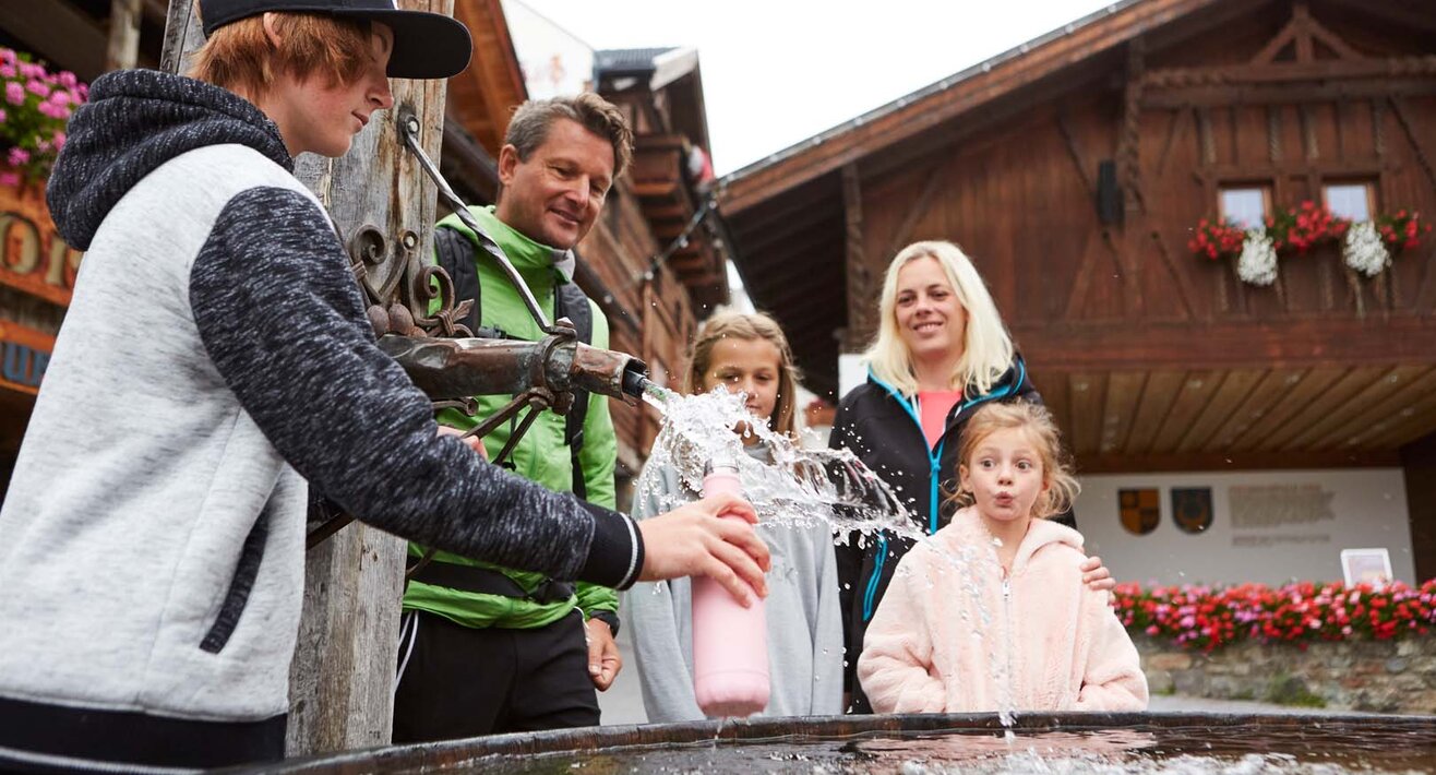 Familie füllt Trinkflasche beim Dorfbrunnen in Fiss auf Familienregion Serfaus-Fiss-Ladis Tirol | © Serfaus-Fiss-Ladis Marketing GmbH | Christian Waldegger