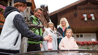 Familie füllt Trinkflasche beim Dorfbrunnen in Fiss auf Familienregion Serfaus-Fiss-Ladis Tirol | © Serfaus-Fiss-Ladis Marketing GmbH | Christian Waldegger