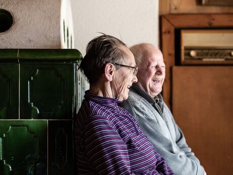 Zwei Ältere aus Ladis sitzen auf einer warmen Ofenbank in einem alten Bauernhaus Beitrag Sun Days Gästemagazin der Region Serfaus-Fiss-Ladis in Tirol | © Serfaus-Fiss-Ladis Marketing GmbH | Rene Raggl