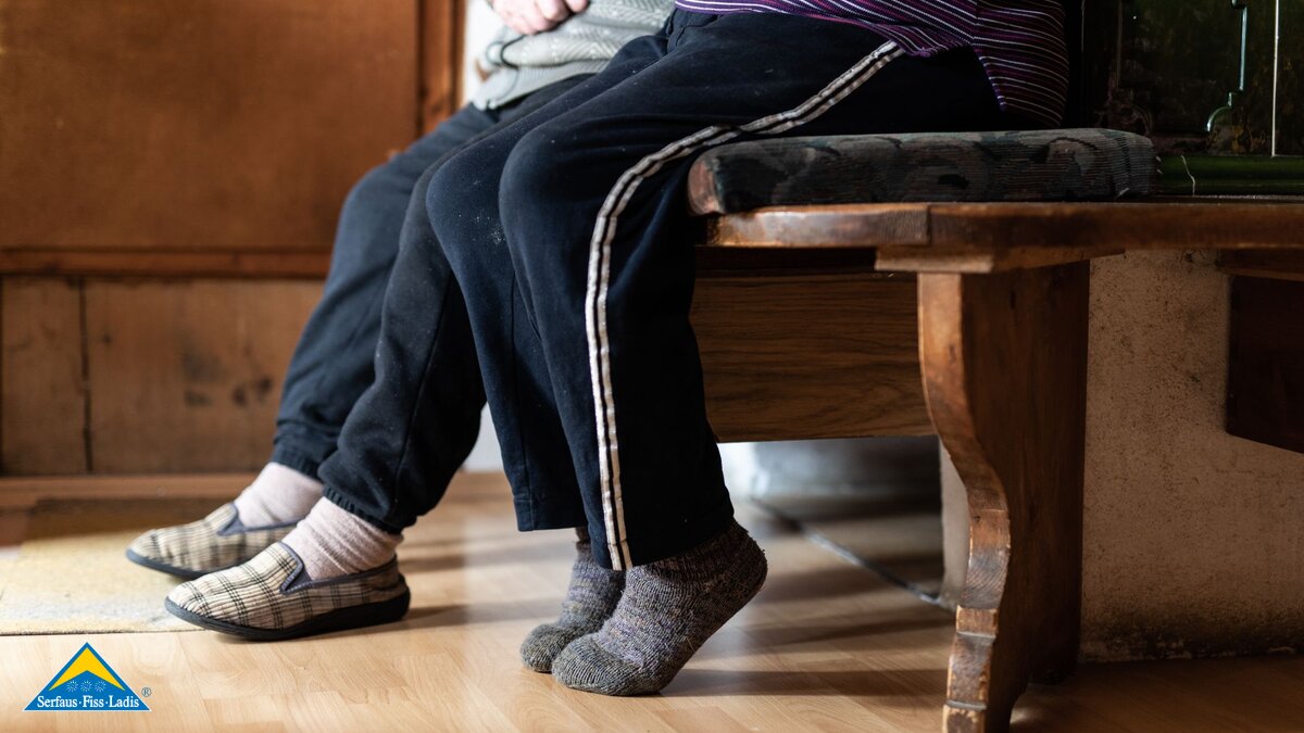 Zwei Personen sitzen auf einer Ofenbank und tragen gewalkte Hausschuhe Blog Serfaus-Fiss-Ladis in Tirol | © Serfaus-Fiss-Ladis Marketing GmbH | Rene Raggl