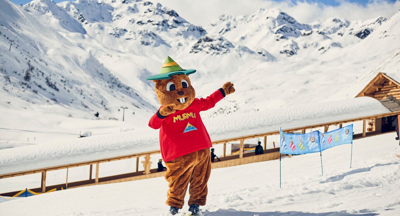 Maskottchen Murmli auf Skiern in der Kinderschneealm im Hintergrund der überdachte Förderbandtunnel Blog Serfaus-Fiss-Ladis in Tirol | © Skischule Serfaus