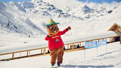 Maskottchen Murmli auf Skiern in der Kinderschneealm im Hintergrund der überdachte Förderbandtunnel Blog Serfaus-Fiss-Ladis in Tirol | © Skischule Serfaus