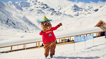 Maskottchen Murmli auf Skiern in der Kinderschneealm im Hintergrund der überdachte Förderbandtunnel Blog Serfaus-Fiss-Ladis in Tirol | © Skischule Serfaus
