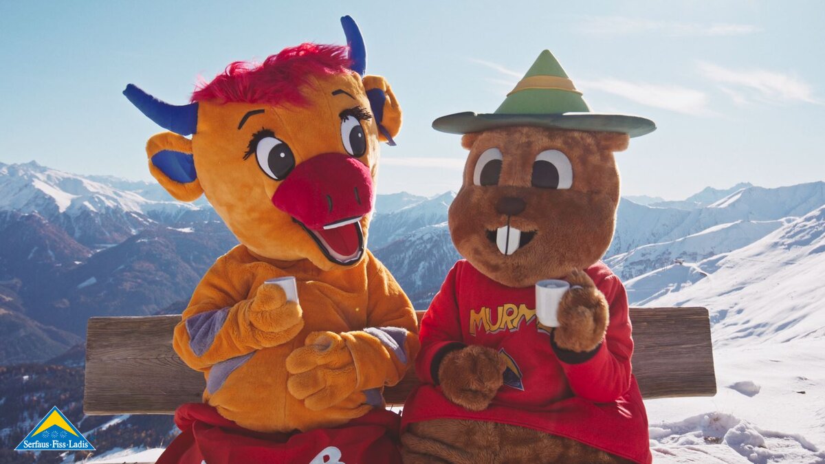 Murmli und Berta im Winter sitzen auf einer Bank am Berg und trinken Kakao Familienregion Serfaus-Fiss-Ladis in Tirol | © Serfaus-Fiss-Ladis Marketing GmbH | Mattias Nigg Films