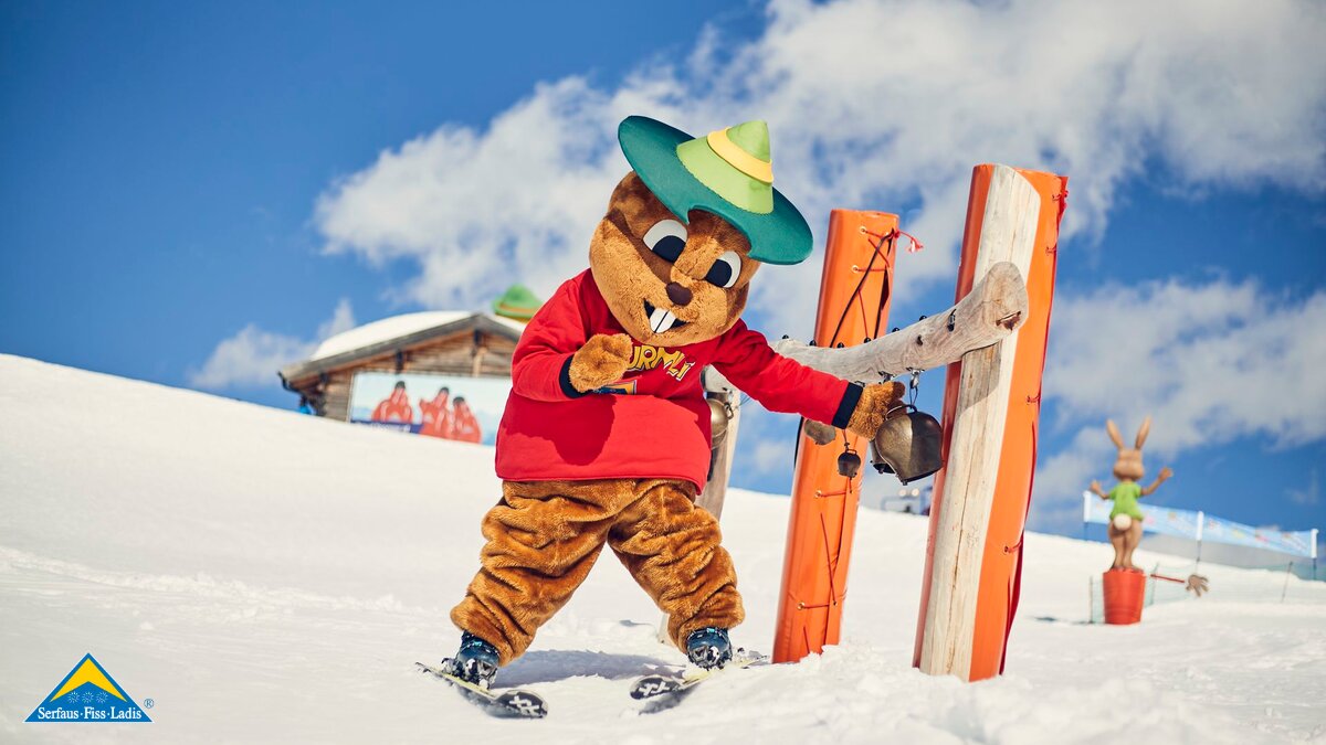 Maskottchen Murmli fährt mit Skiern an einer Spielestation mit Glocken in der Kinderschneealm vorbei Familienregion Serfaus-Fiss-Ladis in Tirol | © Skischule Serfaus