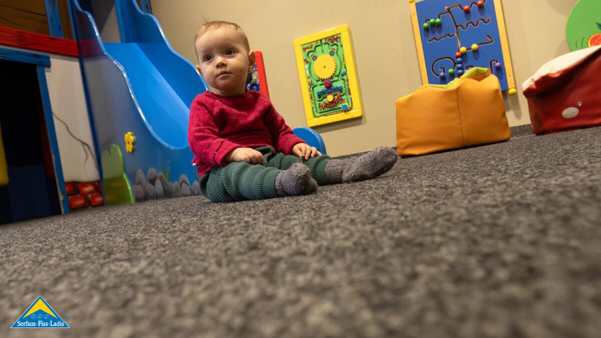 Kleinkind sitzt auf Teppichboden in einem indoor Spielplatz mit Stationen an den Wänden Urlaub mit Kleinkind in Serfaus-Fiss-Ladis in Tirol Tipps | © Serfaus-Fiss-Ladis Marketing GmbH | Andreas Kirschner