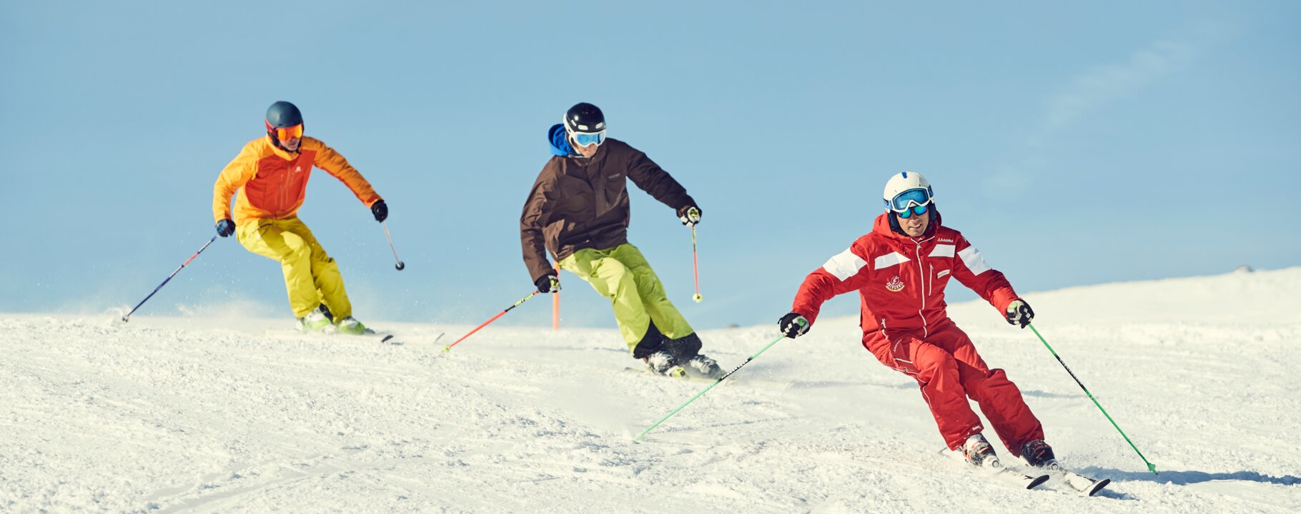 Three skiers, including a ski guide in red ski school gear, are skiing down a snowy slope in a line. The sun is shining, and the sky is clear. The other two skiers are wearing yellow and brown jackets. | © Serfaus-Fiss-Ladis Marketing GmbH | Christian Waldegger